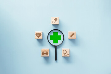 Magnifying glass over green hospital medical icon on blue pastel color background surrounded by wooden block with medical service treatment icon in healthcare and hospitality industry concept.