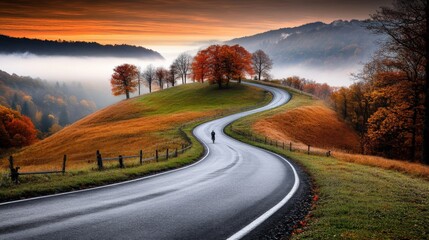 Obraz premium Serene winding road through rolling hills during autumn sunset with colorful trees and misty background