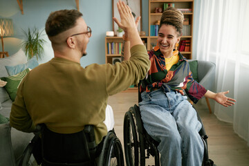 Young adult man with disability giving high five to cheerful woman using wheelchair while smiling...