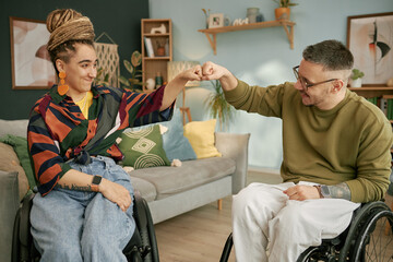 Side view of cheerful man using wheelchair giving fist bump to smiling woman with disability while celebrating success in cozy living room