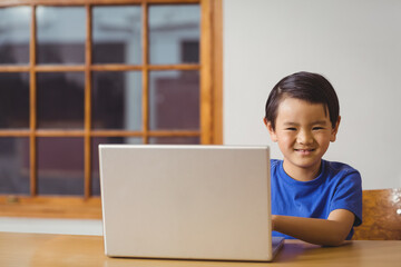 Asian boy sitting at wooden table and chair using laptop near multi-pane window smiling at camera