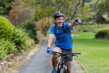 Man riding bicycle with helmet and backpack on paved park path pointing forward with gloved hand