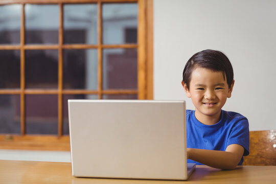 Asian boy sitting at wooden table and chair using laptop near multi-pane window smiling at camera