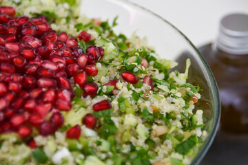 Fresh Tabbouleh salad in a clear glass bowl topped with pomegranate seeds, chopped parsley, bulgur, tomatoes, and mint with olive oil bottle closeup top view of healthy meal