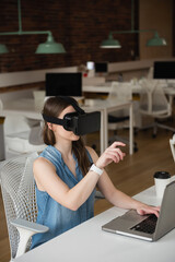 Female employee is wearing VR headset and sitting at white desk, with laptop in open-plan office