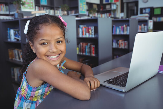 African American girl sitting at table in library using silver laptop by bookshelves, copy space