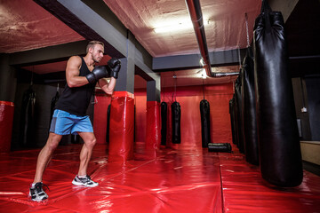 Man practicing boxing in gym basement punching heavy bag with gloves on red mats, copy space