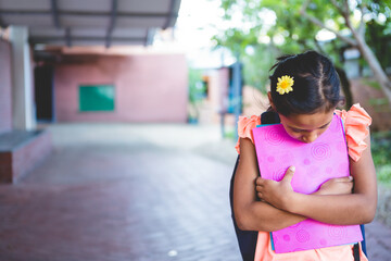 Girl standing clutching pink binder, wearing black backpack on walkway under corridor with trees
