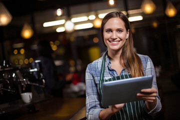 Female barista standing behind wooden counter in cafe using tablet by espresso machine, copy space