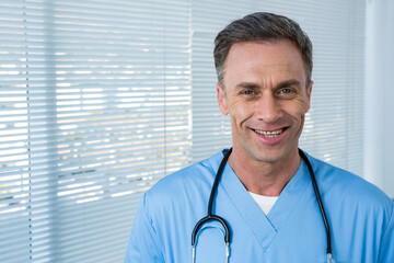 Middle aged doctor standing by window blinds in clinic wearing scrubs and stethoscope, copy space