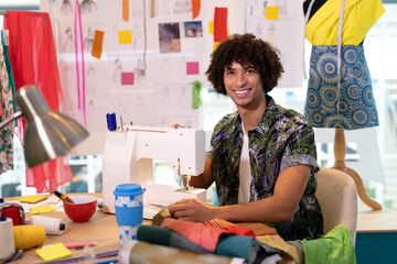 Adult male fashion designer sitting in workshop, using sewing machine with spools of thread