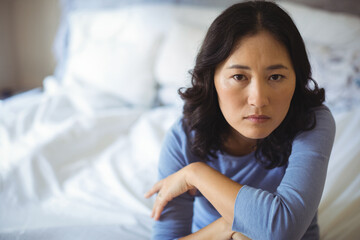Asian woman sitting on bed with white pillows and sheets folding arms and looking serious
