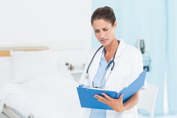 Hospital bed resting with crisp white linens beside vital signs monitor and blue clipboard on table