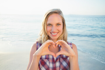 Woman smiling, making heart shape with hands at beach shoreline wearing button-up shirt
