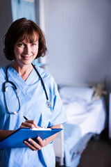 Female clinician writing on clipboard with pen wearing stethoscope beside hospital bed, copy space