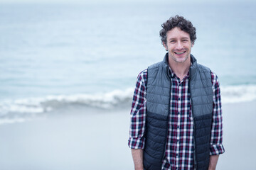 Man in his 40s wearing quilted vest standing on beach with waves rolling, copy space