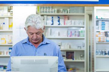 Senior male pharmacist in blue shirt checking white monitor at pharmacy counter and shelves