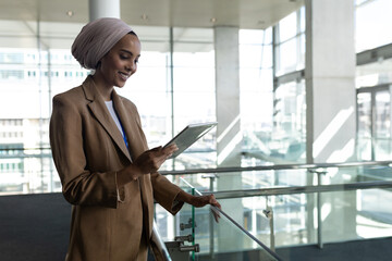 Businesswoman wearing tan blazer and hijab leaning on glass railing and using tablet in atrium