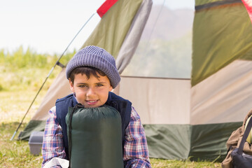 Middle Eastern boy sitting on grass at campsite holding sleeping bag near khaki-red canvas tent