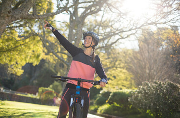 Female cyclist wearing sportswear, helmet and gloves riding bicycle along park path pointing upward
