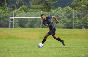 African American male soccer player dribbling ball on field near goalpost wearing jersey and cleats