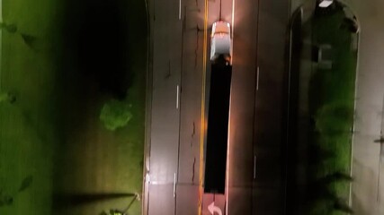Aerial night view of multiple freight trucks lined up in a straight row along a road in an industrial area. The vehicles are parked, waiting for their turn to unload or load cargo at nearby facilities