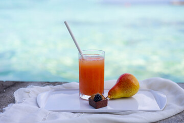 Glass of fresh juice next to pear and cake on tray against background of azure ocean or sea. Beach holiday on exotic island. Travel, vacation content