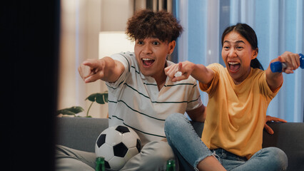 Asian new marriage couple sit on sofa excited cheering watching football sport game on television in living room at home. Young couple sport fans hold football watching online soccer on TV.