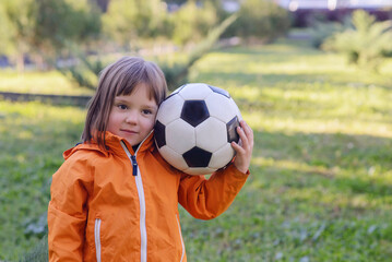 Little toddler girl in orange jacket holding soccer ball on shoulder, standing on green grass in...