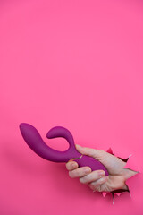Female hand sticking out with raspberry sex toy from pink paper background.