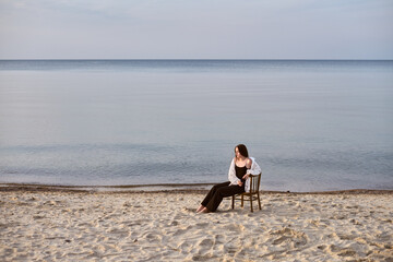 Young woman sits on chair by calm sea on sandy beach, gazing thoughtfully at horizon. Atmosphere is...