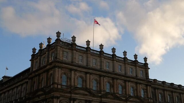 British flag waving in the wind on top of the building in Edinburgh at sunrise. Union Jack flag on a pole brightened with morning sun. Blue sky with white clouds. Historic building in the shade.