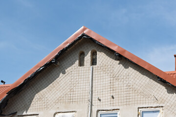 Renovation of a house facade with visible reinforcement mesh, unfinished windows, and a red gable roof against clear blue sky during daylight.