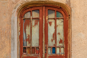Fototapeta premium Vintage arched window with peeling red paint and cracked glass panels in an old textured wall highlights the charm of urban decay and history.