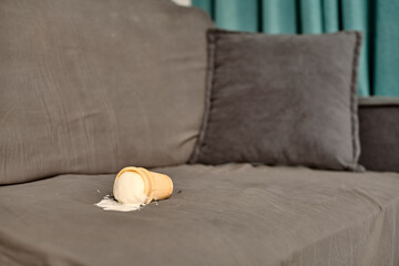 Ice cream cone tipped over on gray sofa, melting onto fabric. Blurred teal curtains in background,...