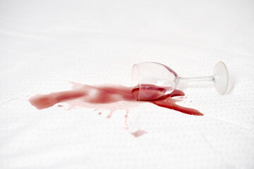 Wine glass toppled over, releasing red wine onto white tablecloth. Stain contrast creates striking...