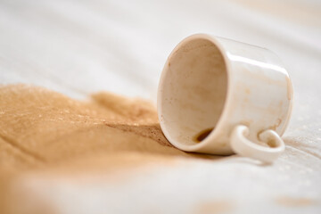 White cup tipped over on fabric, resulting in brown coffee stain spreading outward. Light...