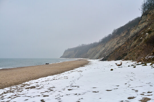 Snow-blanketed beach meets rocky cliffs under cloudy winter sky. Muted tones of shoreline contrast with cold sea. Tranquil, desolate atmosphere fills scene
