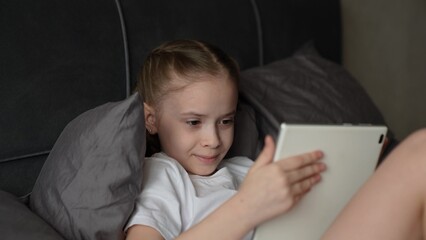 child playing tablet, little girl playing tablet, Girl using tablet while lying in bed relaxing comfortably, Happy child watching cartoon on tablet under gray soft pillows, Young girl smiling at