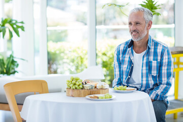 Senior man sitting at table near windows, admiring cheeseboard with grapes, crackers, copy space