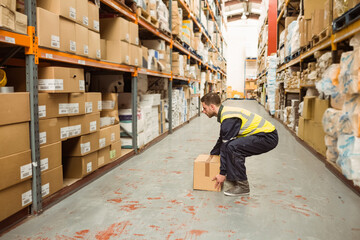Male worker wearing high-visibility vest crouching and lifting cardboard box in storage aisle