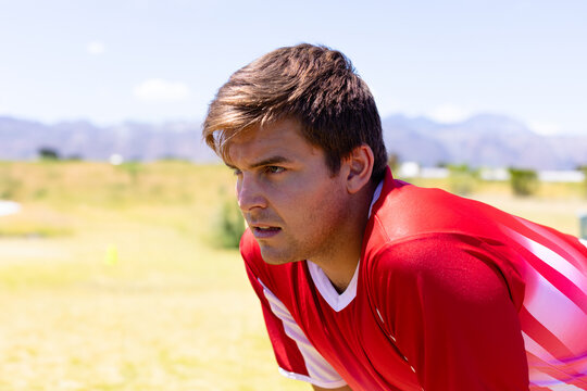 Man leaning forward hands on knees on grass field wearing red jersey, black shorts, copy space