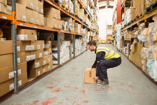 Male worker wearing high-visibility vest crouching and lifting cardboard box in storage aisle - Powered by Adobe