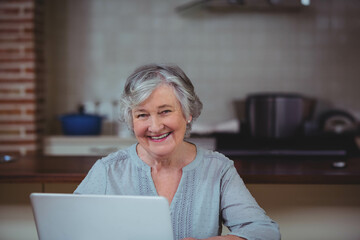 Senior woman typing on laptop at kitchen counter with blue pot and tiled backsplash