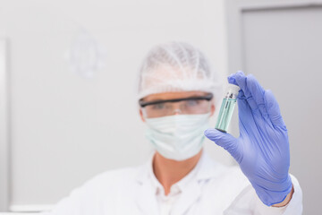 Gloved hand holding small clear vial containing green liquid in sterile lab, near grey cabinet