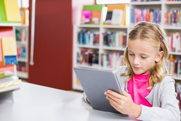 Girl reading tablet at white study table in library reading area, colorful books background