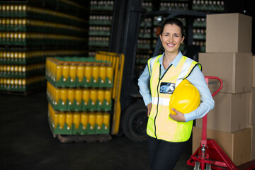 Female warehouse worker wearing safety vest holding hard hat amid beverage crates, copy space
