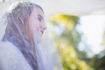 Bride wearing white wedding gown floral veil filtering sunlight glowing blurred greenery at arch