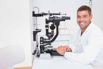 Male optometrist wearing lab coat sitting beside slit lamp biomicroscope at clinic desk, copy space