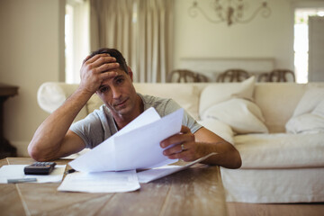 Middle-aged man sitting at home reviewing paperwork using calculator on table and pressing forehead